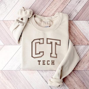 May include: A cream-colored sweatshirt with the text "CT TECH" in brown lettering. The letters are in a bold, blocky font.