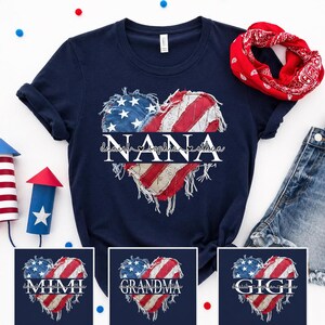 K&ouml;nnte beinhalten: Marineblaues T-Shirt mit einem herzf&ouml;rmigen Design der amerikanischen Flagge, dem Wort "NANA" und zwei Namen. Zus&auml;tzliche Designs zeigen "MIMI", "GRANDMA" und "GIGI" in &auml;hnlichen herzf&ouml;rmigen Flaggen-Designs. Ein rotes Bandana und Jeansshorts sind ebenfalls sichtbar.