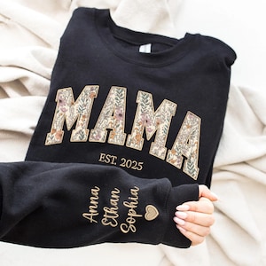 May include: Black sweatshirt with the word "MAMA" in floral letters and the text "EST. 2025" below. The sleeve has the names "Anna", "Ethan", and "Sophia" embroidered in gold thread.
