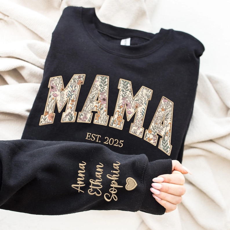May include: Black sweatshirt with the word "MAMA" in floral letters and the text "EST. 2025" below. The sleeve has the names "Anna", "Ethan", and "Sophia" embroidered in gold thread.