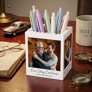May include: A white square pen holder constructed from building blocks featuring a photo of two men embracing on the front. The text "Our Story Continues" and "HAPPY FATHER'S DAY" is printed below the photo. Several pastel-colored pens are inside.