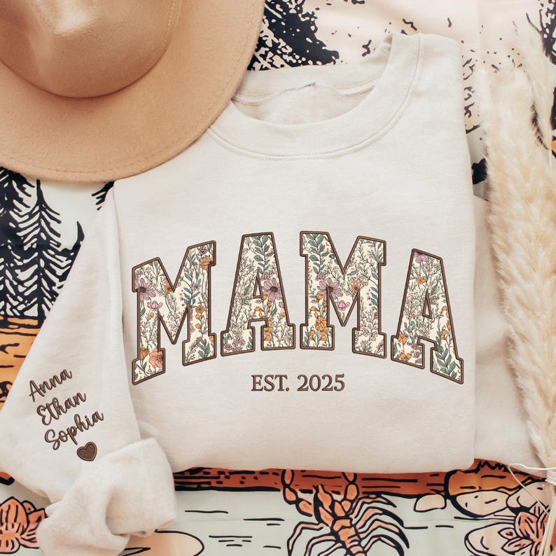 May include: A cream-colored sweatshirt with the word "MAMA" in large, floral letters. The letters are outlined in brown and filled with a floral pattern. The text "EST. 2025" is printed below the word "MAMA".
