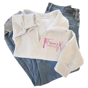 May include: A light gray quarter-zip sweatshirt with the text "Fave's Hairstylist" in pink, along with a comb and scissors graphic. The sweatshirt is paired with blue denim jeans.
