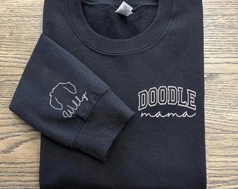 May include: A black sweatshirt with the words "DOODLE mama" in white lettering. The left sleeve features a doodle dog outline and the name "Willy" in white script. The sweatshirt is folded neatly on a wooden surface.