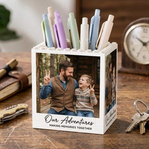 May include: A white square desk organizer constructed from building blocks, featuring a photo of a man and a child on the front. The text "Our Adventures MAKING MEMORIES TOGETHER" is printed below the photo. Several pastel-colored pens are stored inside.