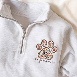 May include: A light gray quarter-zip sweatshirt featuring a floral paw print design with the text "dog mama". The paw print has a brown border and is filled with colorful flowers. The sweatshirt has a silver zipper.