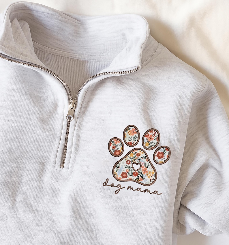 May include: A light gray quarter-zip sweatshirt featuring a floral paw print design with the text "dog mama". The paw print has a brown border and is filled with colorful flowers. The sweatshirt has a silver zipper.