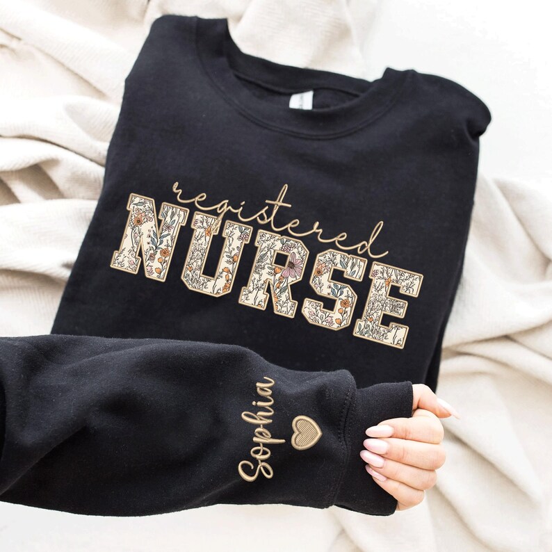 May include: Black sweatshirt with the text "registered NURSE" in floral lettering. The word "NURSE" is surrounded by a floral pattern. The sweatshirt has a small heart and the name "Sophia" embroidered on the sleeve.