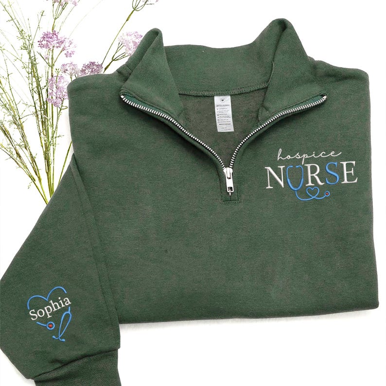 May include: A dark green quarter-zip sweatshirt with the words "hospice NURSE" embroidered in white and blue. The sleeve features a blue heart with the name "Sophia" and a stethoscope design. The sweatshirt has a silver zipper.