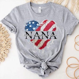 K&ouml;nnte beinhalten: Ein graues T-Shirt mit einem herzf&ouml;rmigen Design, das die Sterne und Streifen der amerikanischen Flagge zeigt. Das Wort "NANA" ist &uuml;ber dem Herzen aufgedruckt, darunter die Namen Daniel, Sophia und Ethan.