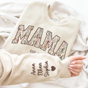 May include: A cream colored sweatshirt with the word "MAMA" in large, floral letters. The sweatshirt has a brown heart and the names "Anna", "Ethan", and "Sophia" written on the sleeve.
