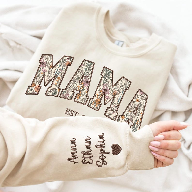 May include: A cream colored sweatshirt with the word "MAMA" in large, floral letters. The sweatshirt has a brown heart and the names "Anna", "Ethan", and "Sophia" written on the sleeve.