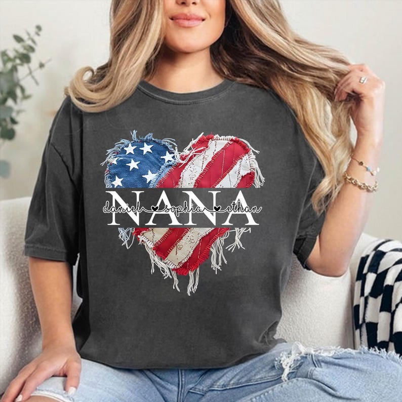 K&ouml;nnte beinhalten: Dunkelgraues T-Shirt mit einem herzf&ouml;rmigen Design der amerikanischen Flagge. Das Wort "NANA" ist prominent dargestellt, mit den Namen "Daniela, Sophia, Ethan" darunter. Das Design hat einen Used-Look.