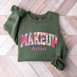 May include: A green sweatshirt with the word "MAKEUP" in pink letters with a floral pattern. The word "Artist" is printed below the word "MAKEUP".