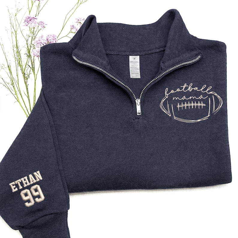 May include: A dark navy blue quarter-zip sweatshirt featuring a white embroidered football graphic with the text "football mama" on the chest. The sleeve is embroidered with the name "ETHAN" above the number "99" in a matching white thread.