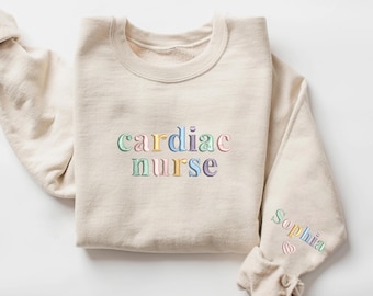 May include: A beige sweatshirt with the words "cardiac nurse" embroidered in colorful letters. The left sleeve has the name "Sophia" embroidered in a heart shape.