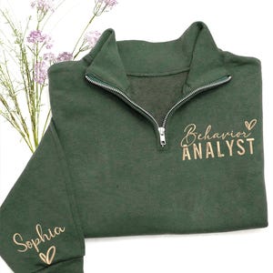 May include: A dark green quarter-zip sweatshirt with the words "Behavior Analyst" and the name "Sophia" in a cursive font. The sweatshirt has a silver zipper and a small heart symbol next to the text. The background is white with some purple flowers.