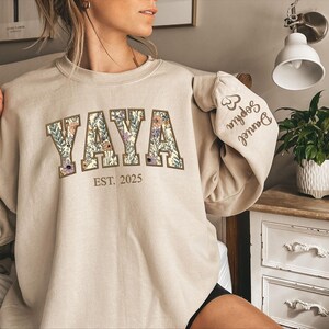 May include: Beige sweatshirt with the word "YAYA" in large floral letters. Below the word "YAYA" is the text "EST. 2025". The sleeve has the names "Daniel" and "Sophia" printed on it. The sweatshirt is a neutral color.