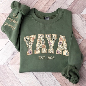 May include: Green sweatshirt with the word "YAYA" in a floral design. The text "EST. 2025" is below the word "YAYA". The left sleeve has the names "Sophia" and "Daniel" embroidered on it.