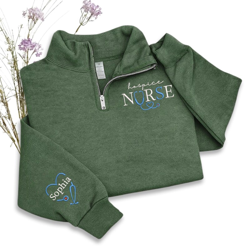 May include: A green quarter-zip sweatshirt with the embroidered text "hospice NURSE" and a stethoscope design. The left sleeve features a heart with the name "Sophia" and a stethoscope. The sweatshirt is made of a soft material.