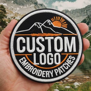 May include: Round black embroidered patch with white and orange text and design. The patch features the words "CUSTOM LOGO EMBROIDERY PATCHES" in white and orange. A mountain range and rising sun are also featured.