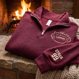 May include: A maroon quarter-zip sweatshirt featuring gold embroidery on the chest that reads football mom with a football graphic. The sleeve is embroidered with the name ETHAN and the number 99 in gold thread. The garment has a silver zipper.