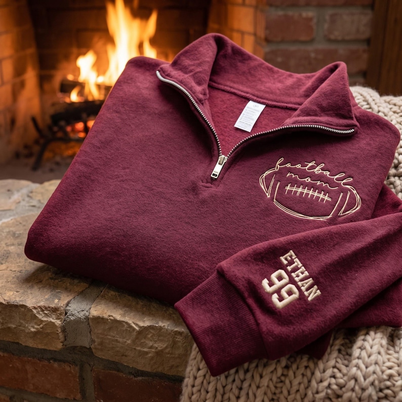 May include: A maroon quarter-zip sweatshirt featuring gold embroidery on the chest that reads football mom with a football graphic. The sleeve is embroidered with the name ETHAN and the number 99 in gold thread. The garment has a silver zipper.