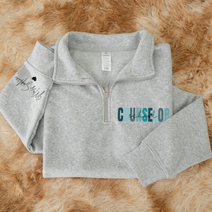 May include: A light gray quarter-zip sweatshirt with the word "COUNSELOR" in teal and blue lettering. The sleeve has the text "Mrs. Sophia" in script with a small black heart. The sweatshirt is made of a soft material.