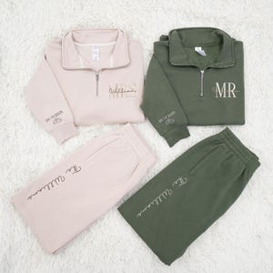 May include: Matching set of a light pink sweatshirt and sweatpants, and an olive green sweatshirt and sweatpants. The sweatshirts have a quarter-zip collar and embroidered text that says "MRS Williams" and "MR" respectively. The sweatpants have the embroidered text "The Williams".