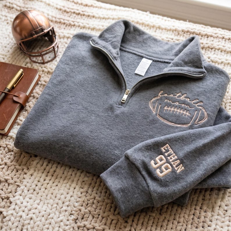 May include: A charcoal gray quarter-zip pullover featuring rose gold embroidery on the chest with a football graphic and the text "football mom". The sleeve is embroidered with the name "ETHAN" and the number "99" in matching rose gold thread.