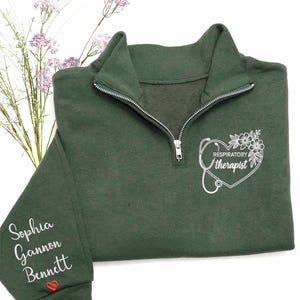 May include: A dark green quarter-zip sweatshirt with white embroidered text and graphics. The right chest features a heart-shaped design with a stethoscope and the words "Respiratory Therapist." The left sleeve has three names embroidered in white, with a red heart below.