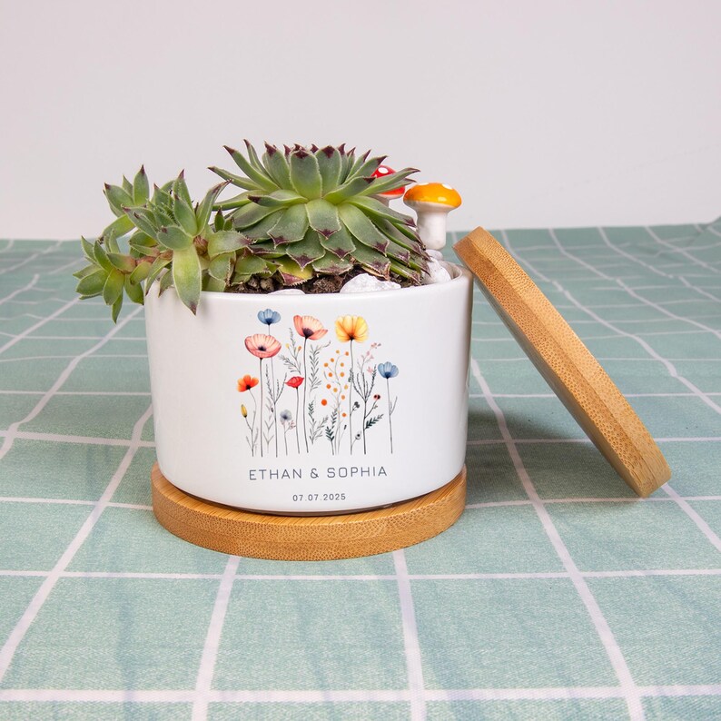 May include: White ceramic planter with a floral design and a wooden base. The planter contains a succulent plant, white pebbles, and a small mushroom figurine. The names "ETHAN & SOPHIA" and the date "07.07.2025" are printed on the planter.