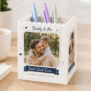 May include: White pen holder with a photo of a father and child, the text "Daddy & Me", "Best Dad Ever", and "Happy Father's Day". The holder has a brick-like design and contains blue, purple, and green pens.