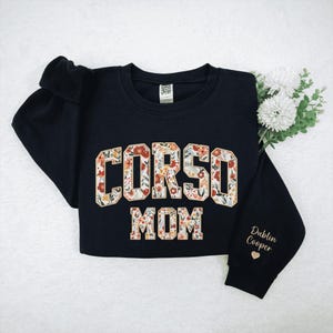 Op de afbeelding: Zwart sweatshirt met de tekst "CORSO MOM" in een bloemenpatroon. De mouw is geborduurd met "Dublin Cooper" in goud, vergezeld van een klein hartje. Het kledingstuk ligt plat op een witte achtergrond.