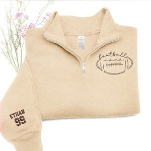 May include: A beige quarter-zip sweatshirt featuring a dark brown graphic of a football with the text "football mama" on the chest. The sleeve is embroidered with the name "ETHAN" above the number "99" in dark brown thread.
