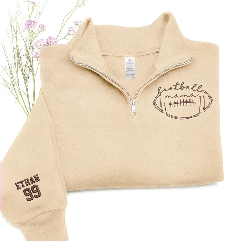 May include: A beige quarter-zip sweatshirt featuring a dark brown graphic of a football with the text "football mama" on the chest. The sleeve is embroidered with the name "ETHAN" above the number "99" in dark brown thread.