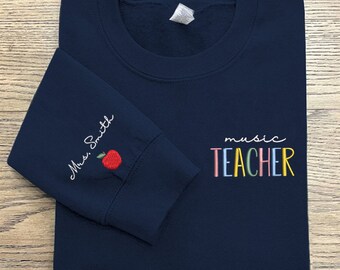 May include: Navy blue sweatshirt with the text "music TEACHER" in rainbow colors on the front and "Miss Smith" with a red apple on the left sleeve.
