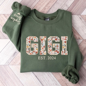 May include: An olive green sweatshirt with the word "GIGI" in a floral design across the chest. Below the word "GIGI" is the text "EST. 2024". The left sleeve has the embroidered name "Sophia Louis".
