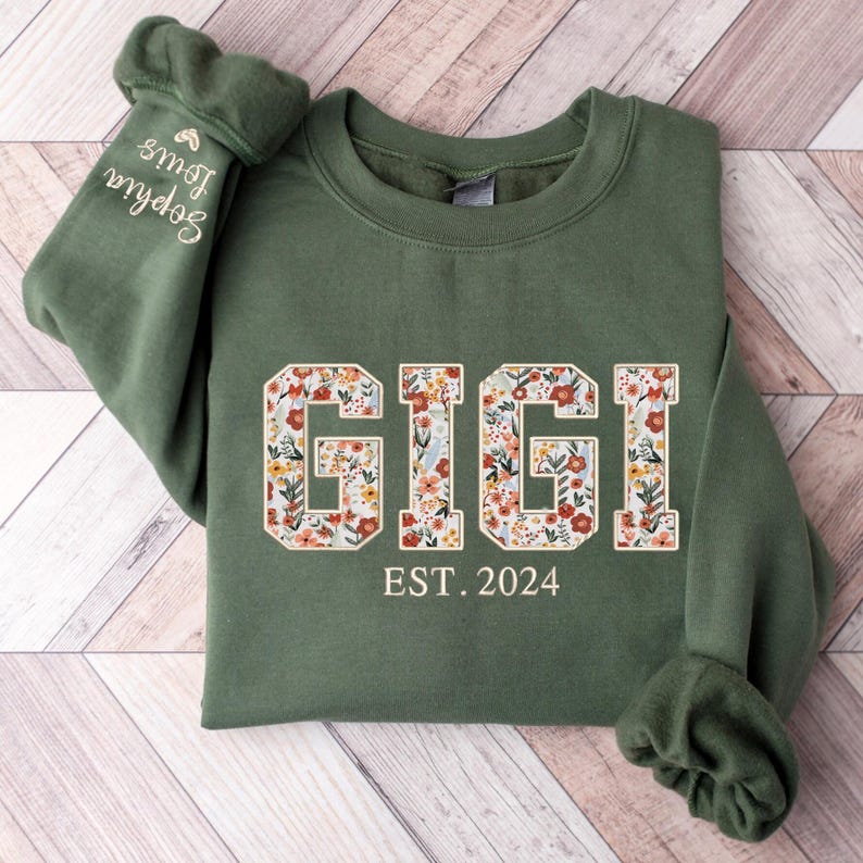 May include: An olive green sweatshirt with the word "GIGI" in a floral design across the chest. Below the word "GIGI" is the text "EST. 2024". The left sleeve has the embroidered name "Sophia Louis".