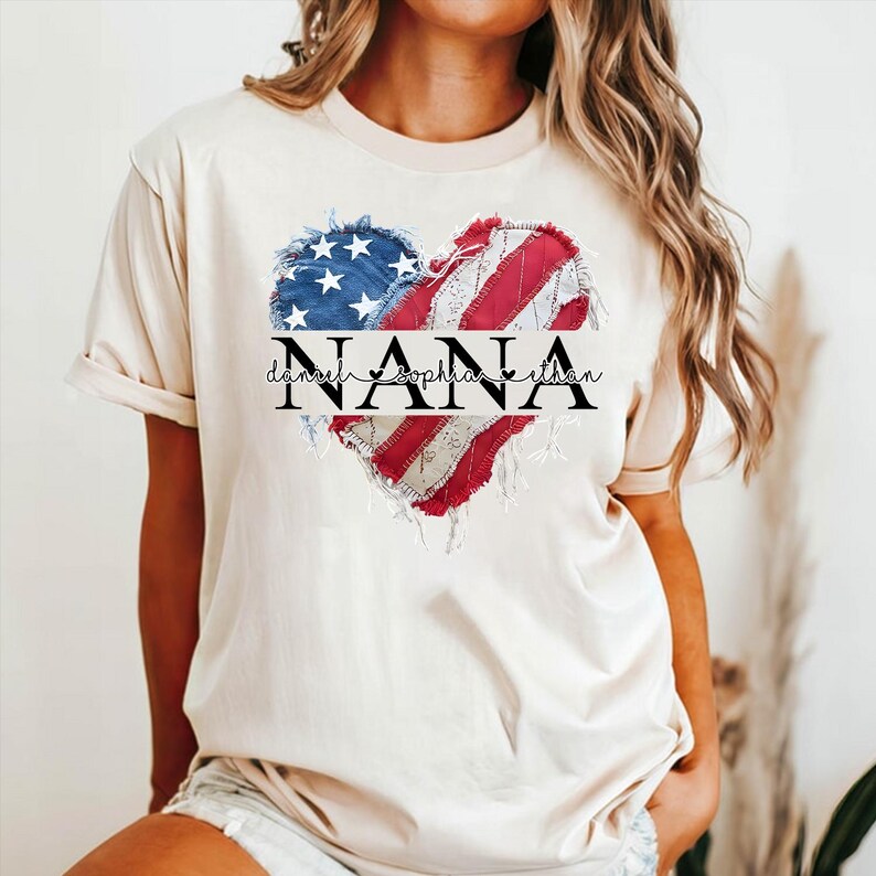 K&ouml;nnte beinhalten: Cremefarbenes T-Shirt mit einem herzf&ouml;rmigen Design, das an die amerikanische Flagge erinnert. Das Design enth&auml;lt das Wort "NANA" in fetten schwarzen Buchstaben, mit den Namen "Daniel, Sophia, Ethan" darunter. Das Herz hat einen Used-Look.