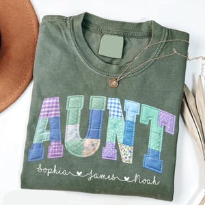 May include: An olive green t-shirt with the word "AUNT" in patchwork letters. The letters are in various patterns and colors, including purple, blue, and floral prints. Below the word are the names "Sophia", "James", and "Noah". A gold necklace is draped across the shirt.