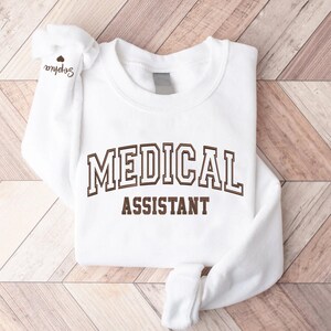 May include: White sweatshirt with brown text that reads "MEDICAL ASSISTANT" in a collegiate font.