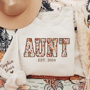 May include: A white sweatshirt with the word "AUNT" in brown letters with a floral pattern. The text "EST. 2024" is below the word "AUNT".