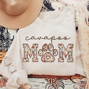 May include: A cream-colored sweatshirt with the text "cavapoo MOM" in brown lettering. The letters are decorated with a floral pattern and a paw print in the center. The sweatshirt has a crew neck and long sleeves.
