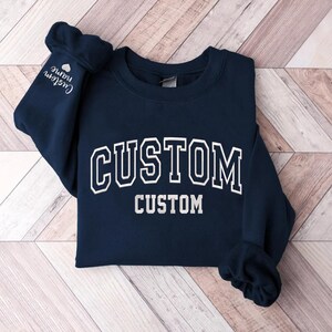 May include: Navy blue sweatshirt with white text that reads "CUSTOM" in a large, bold font and "CUSTOM" in a smaller font below. The sweatshirt has a crew neck and long sleeves.