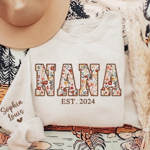 K&ouml;nnte beinhalten: Ein cremefarbener Pullover mit dem Wort "NANA" in gro&szlig;en braunen Buchstaben. Die Buchstaben sind mit einem floralen Muster in Rosa-, Gelb- und Gr&uuml;nt&ouml;nen gef&uuml;llt. Der Text "EST. 2024" ist unter dem Wort "NANA" gedruckt.