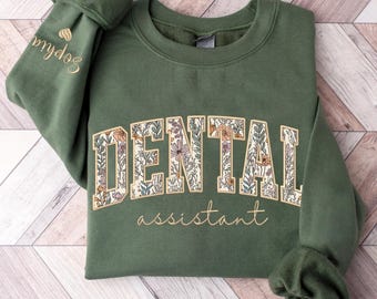 May include: A green sweatshirt with the words "DENTAL assistant" in a floral design across the front. The sleeve has the name "Sophia" embroidered in gold. The sweatshirt is folded on a wooden surface.