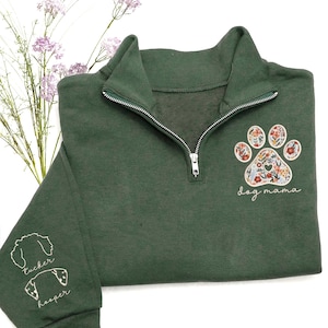 May include: Dark green quarter-zip sweatshirt with a floral paw print design and the words "dog mama." The sleeve features embroidered dog silhouettes with the names "Tucker" and "Hooper." The sweatshirt has a silver zipper and a high collar.