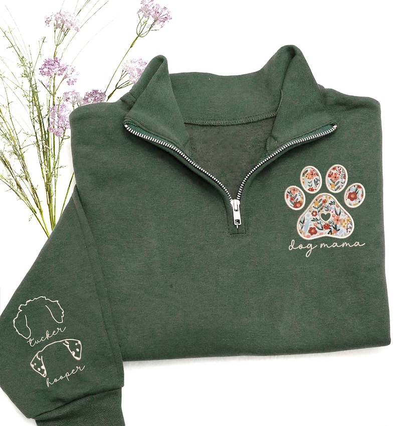 May include: Dark green quarter-zip sweatshirt with a floral paw print design and the words "dog mama." The sleeve features embroidered dog silhouettes with the names "Tucker" and "Hooper." The sweatshirt has a silver zipper and a high collar.