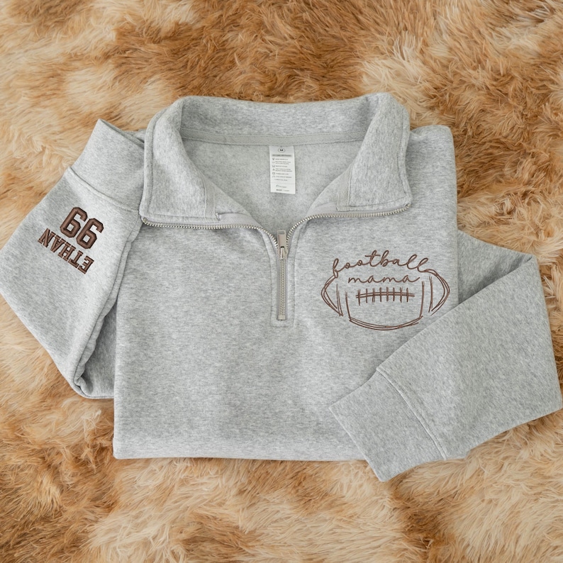 May include: A light gray quarter-zip pullover sweatshirt featuring a brown embroidered football graphic with the text "football mama" on the front and the number "66" and the name "ETHAN" embroidered in brown on the left sleeve.